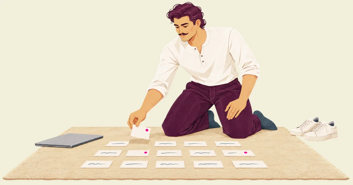 Ivo kneels in socks on a rug laid out with rows of index cards, holding one card above the rows as he decides where it goes, a closed laptop at one edge of the rug and his trainers beside it.